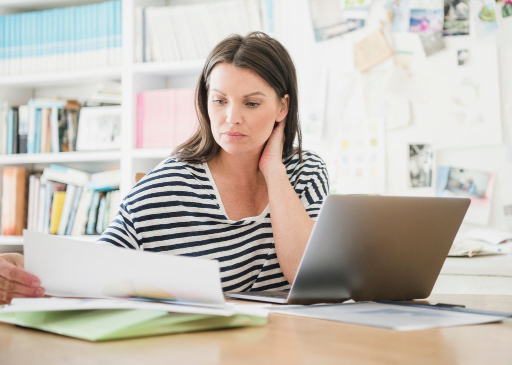 Mid adult businesswoman in home office using computer and looking at paperworkSerious looking woman working from home with laptop, reading document