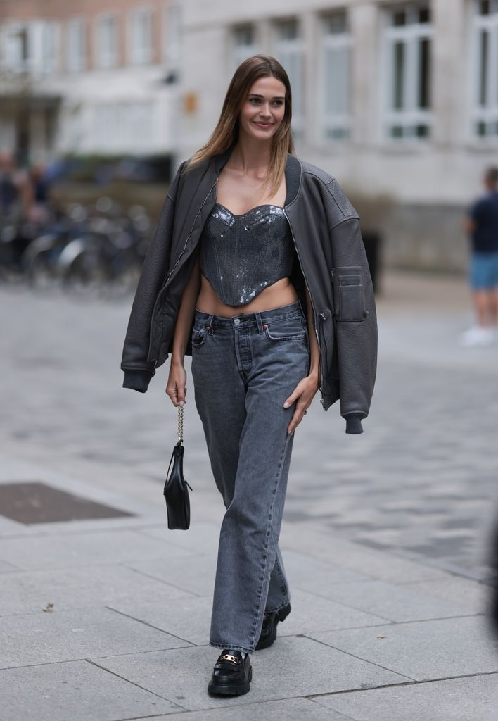 A Fashion Week guest is seen wearing a grey bomber jacket, underneath a grey sequin corset top, grey jeans with straight leg, a black leather bag and black leather loafers with golden chain detail during London Fashion Week September 2023 before the David Koma Show