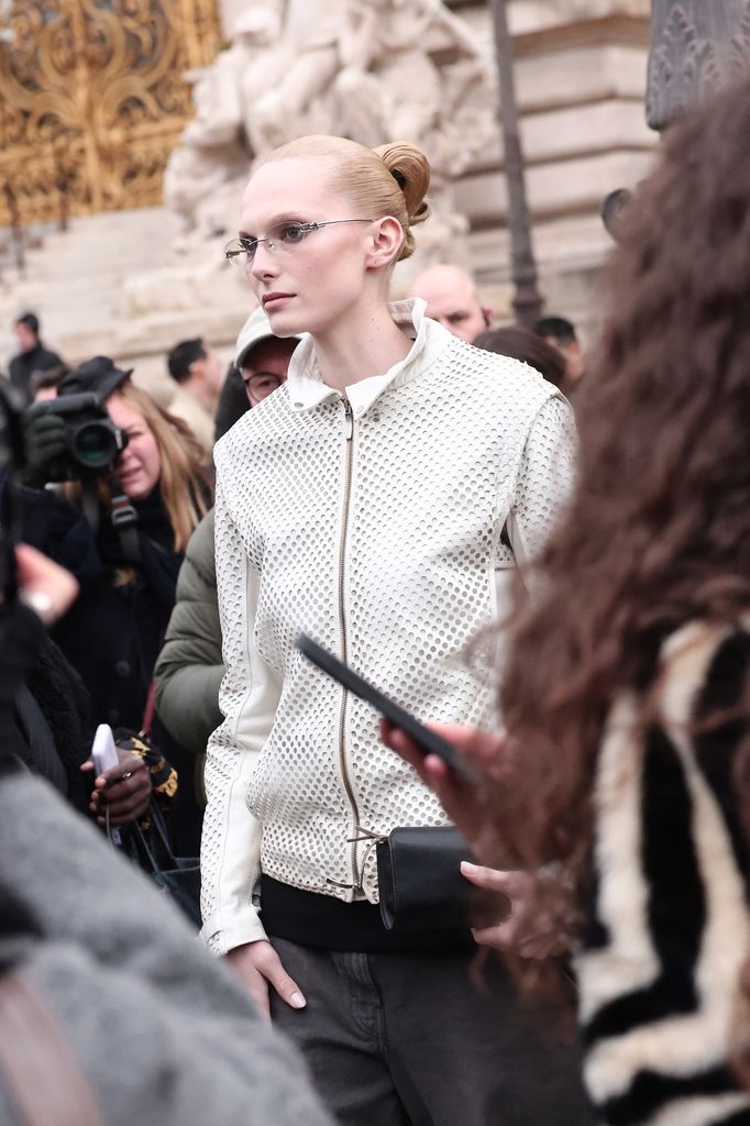 Alex Consani wears a grey jacket with no collar and wears her hair in a slickback bun