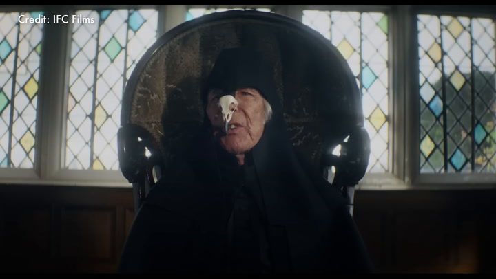 A man with a mask sits on a chair in front of a stained glass window