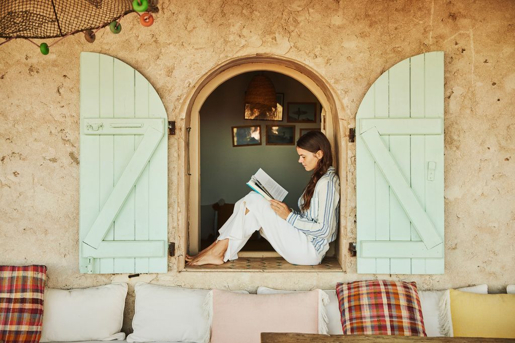 Wide shot of woman sitting in window while relaxing and reading book at tropical villa during vacation in Morocco
