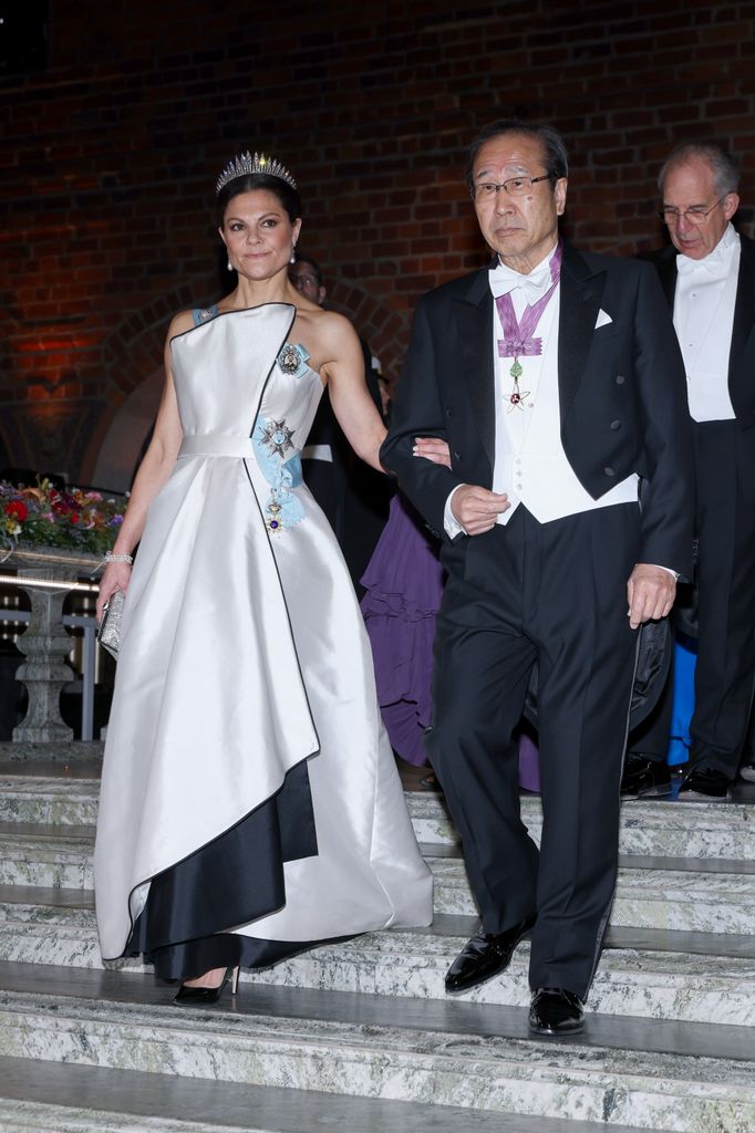 Crown Princess Victoria of Sweden in white gown and tiara holding arm of Susumu Kitagawa, the Japanese chemist, descending stairs