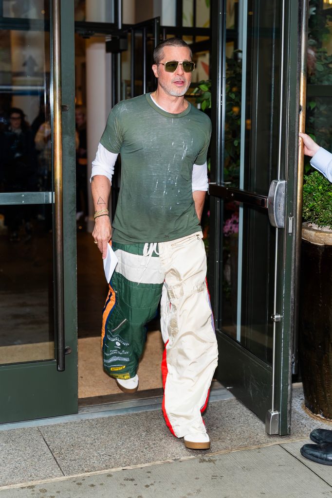 Brad Pitt wears a khaki short sleeve t-shirt over a white long sleeve top, paired with trousers - one leg is beige and the other khaki