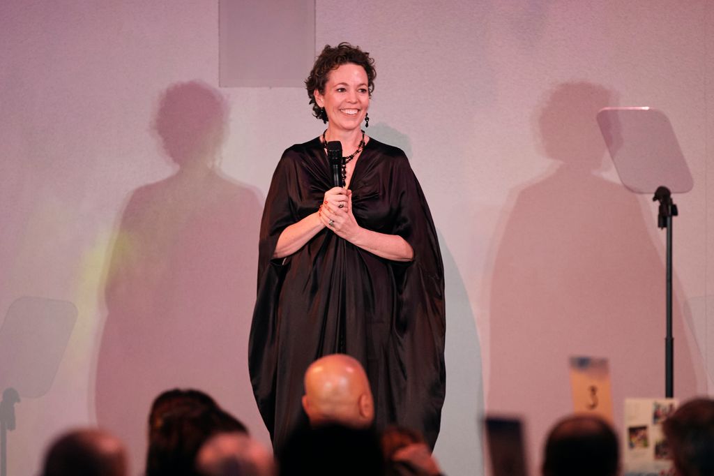 Olivia Colman in a brown caped dress holding a microphone on stage