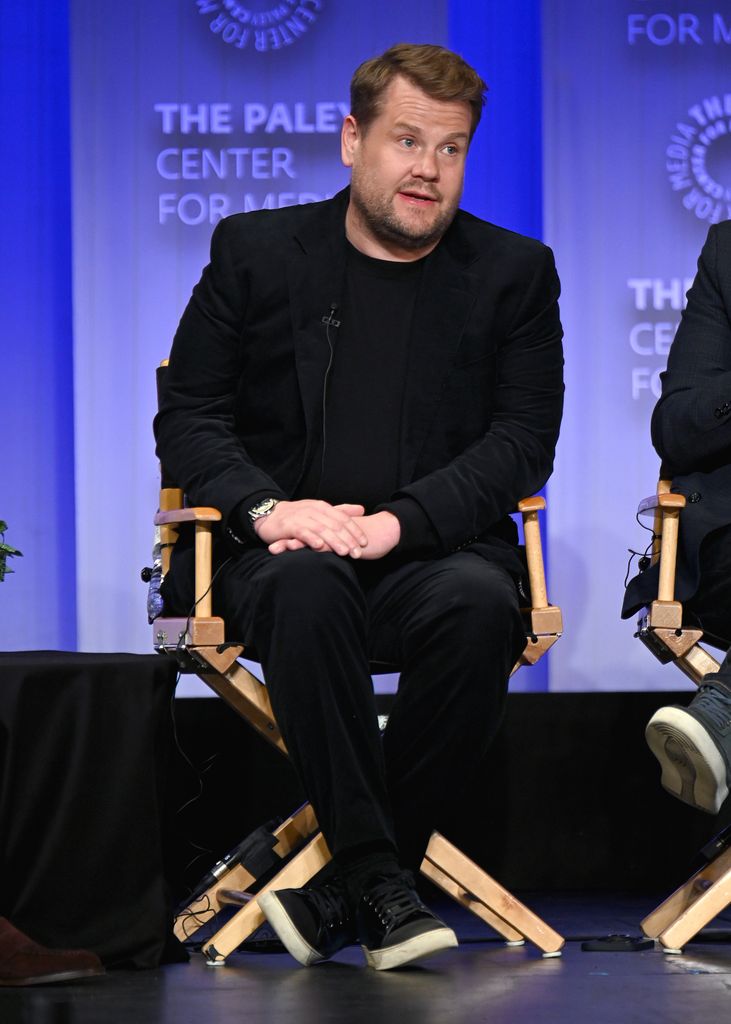 James Corden wearing a black suit sitting on a chair while being interviewed 
