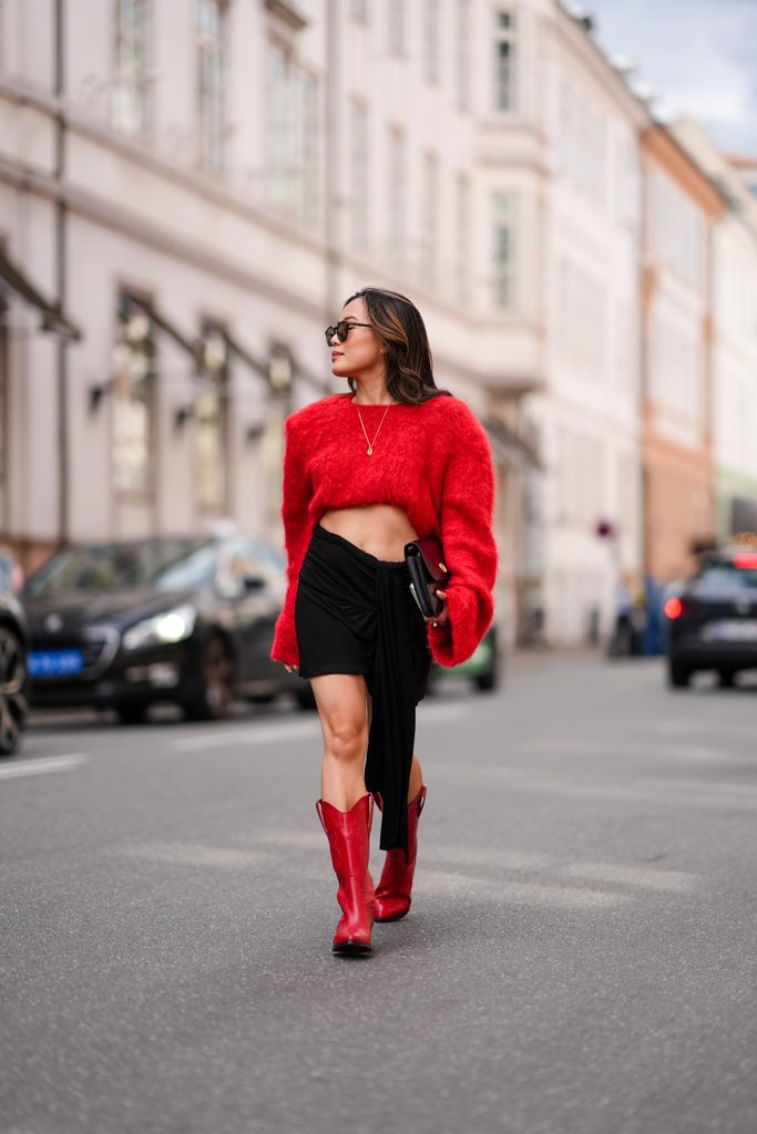 : A guest wears black sunglasses, gold necklace, bright red cropped long sleeve sweater, shiny black leather handbag, black mini skirt, shiny red cowboy boots leather shoes, outside Gestuz, during the Copenhagen Fashion Week Spring/Summer 2024-2025 on August 8, 2024 in Copenhagen, Denmark. (Photo by Edward Berthelot/Getty Images)