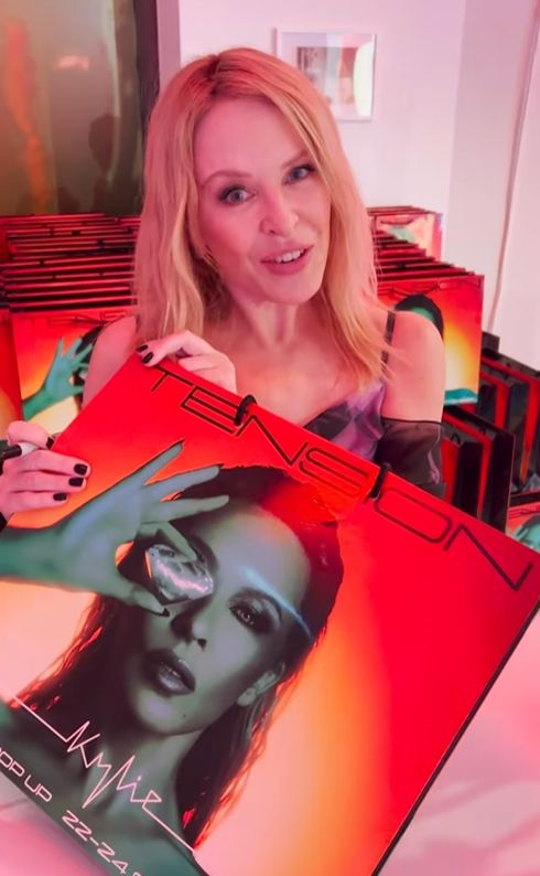 Kylie Minogue in dress stood behind a bag with Tension art