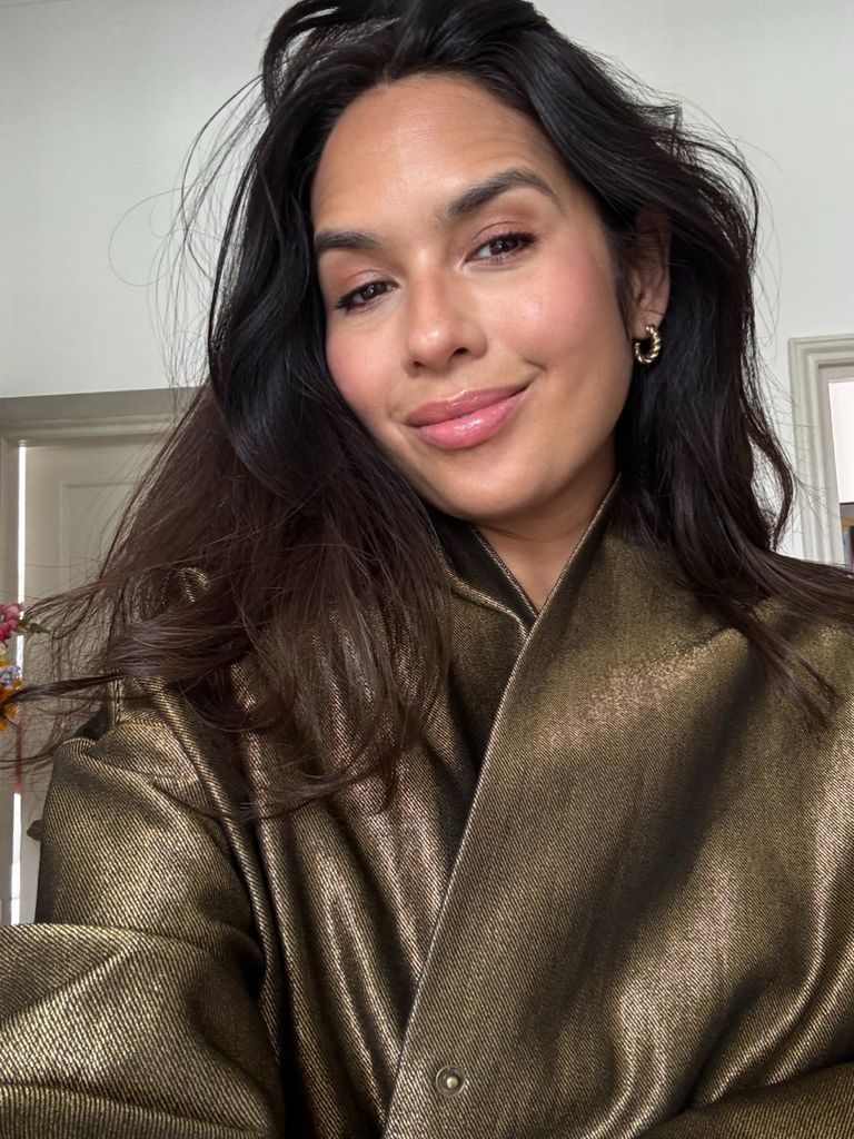 Nieve Tierney smiles softly at the camera in a golden metallic jacket with loose, flowing hair and minimal makeup, radiating natural confidence.