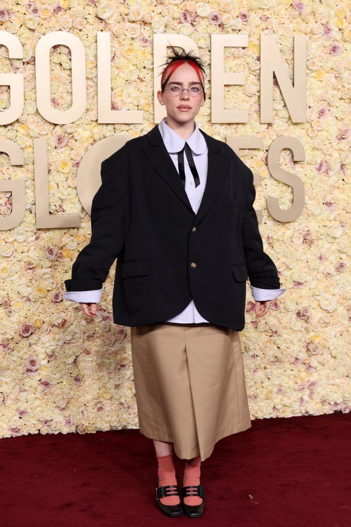 Billie Eilish sports an oversized black Gucci blazer, a khaki skirt, and red socks with chunky Mary Janes. She wears glasses and a red headband, looking quirky yet fashion-forward.