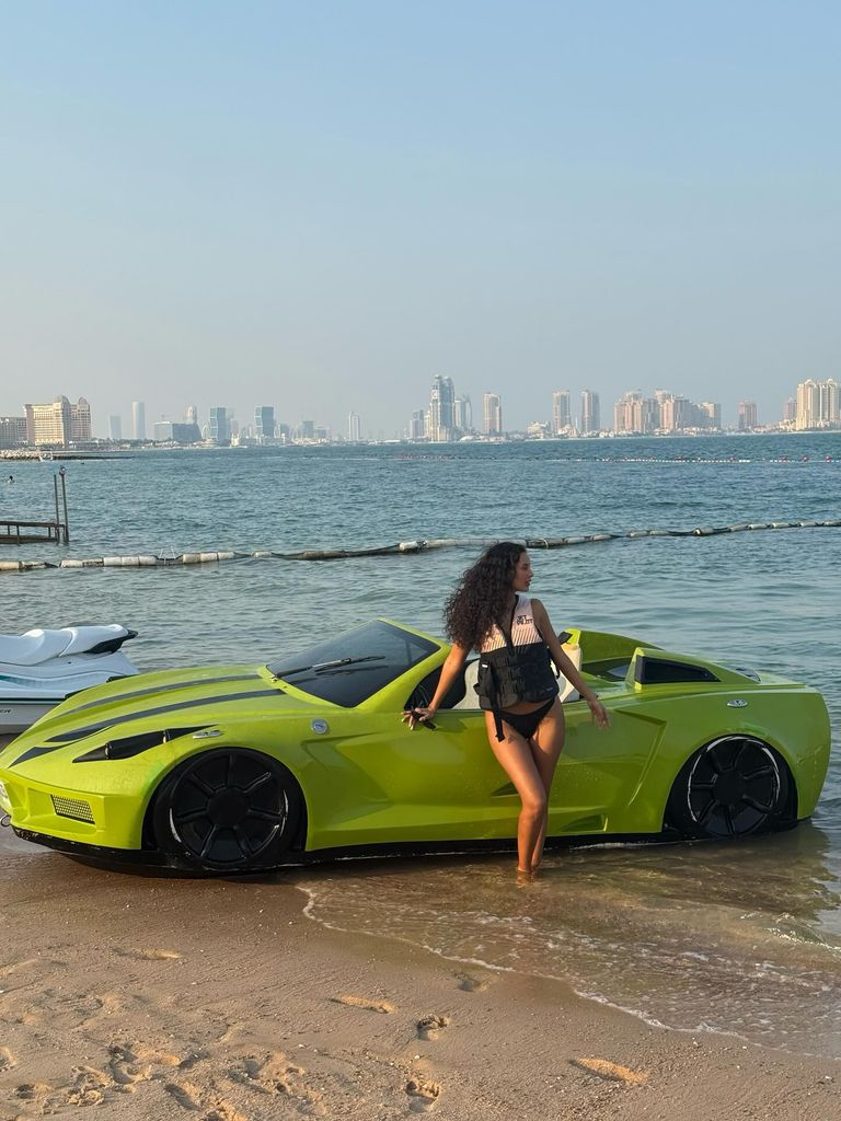 Maya Jama wears a black bikini and life jacket and poses next to a green sports car that is parked on the beach, with the city skyline in view