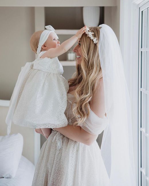 stacey wears a wedding dress and veil as she lifts her daughter who is wearing a white flower girl outfit and reaches out to gently touch Staceys flower garland as they both smile