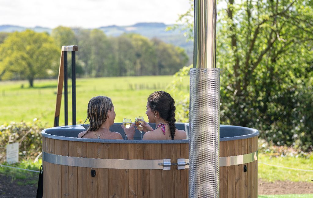 Two women in an outdoor hot tub holding glasses of wine against a backdrop of green fields and trees