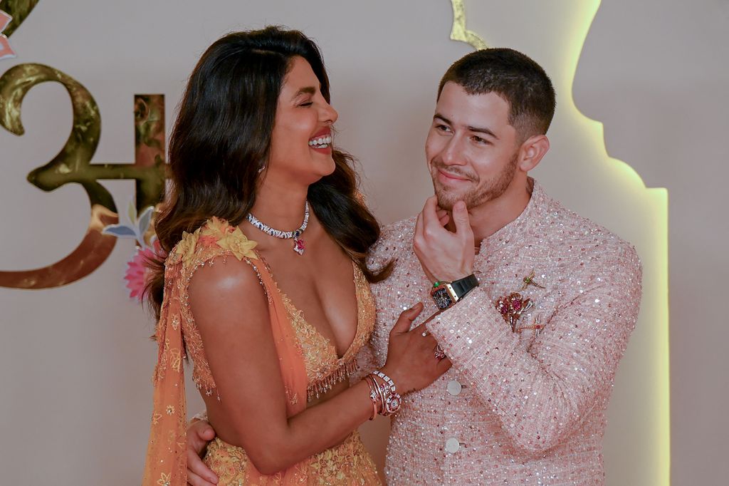 Priyanka Chopra Jonas (L) and her husband American singer-songwriter and actor Nick Jonas pose for a photo, with her laughing while he has his arm around her. She is wearing a sequin orange-toned dress and he wears a faded pink sparkly suit. 