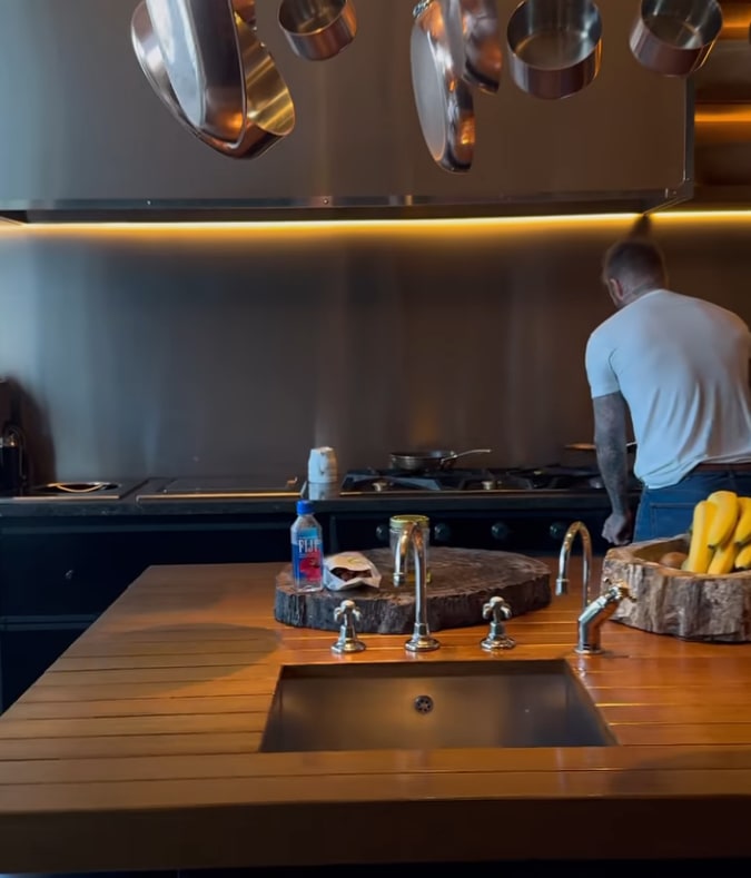 David Beckham frying a crepe on his hob, the foreground shows their kitchen island with sink and chopping boards