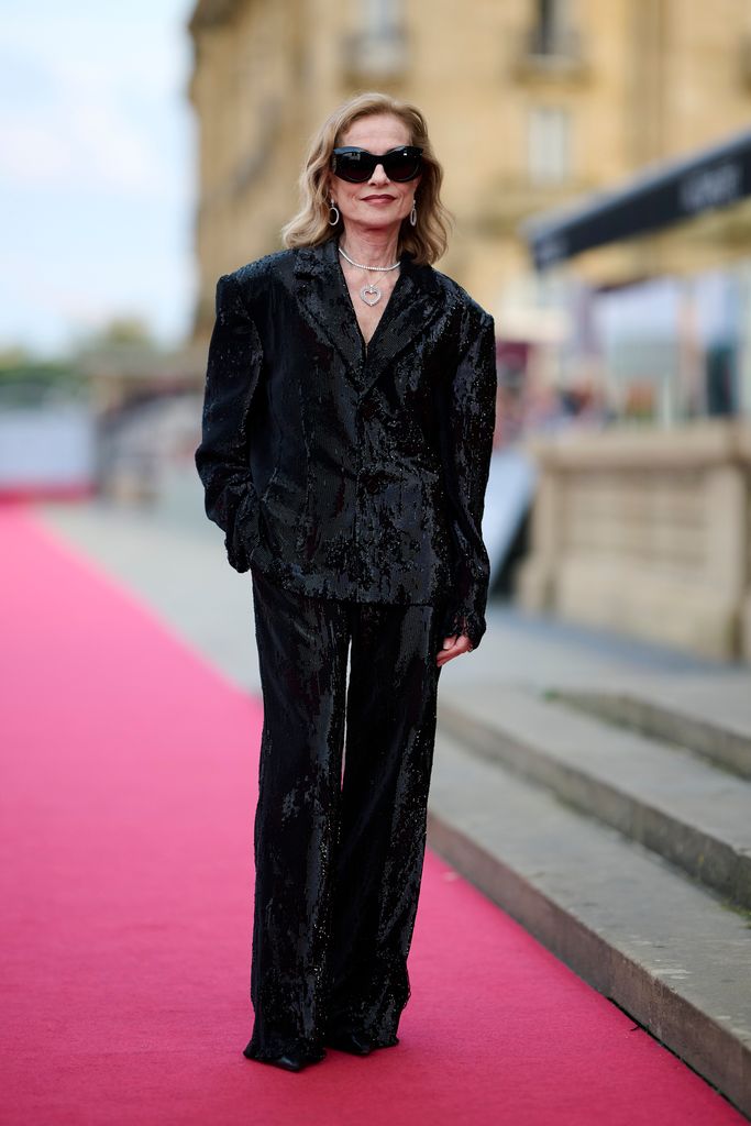 Isabelle Huppert in a shiny black suit with a boxy silhouette, accessorized with oversized sunglasses and a silver necklace. The background is a red carpet event in a historic urban setting.