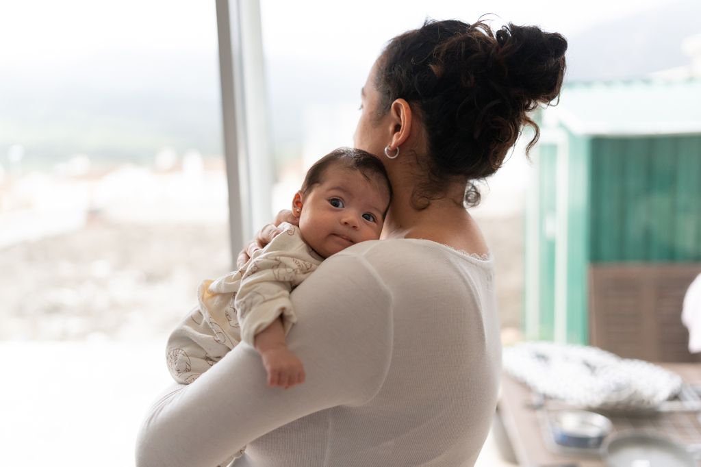 La mère regarde par la fenêtre tout en tenant le bébé dans ses bras. Le bébé regarde la caméra.
