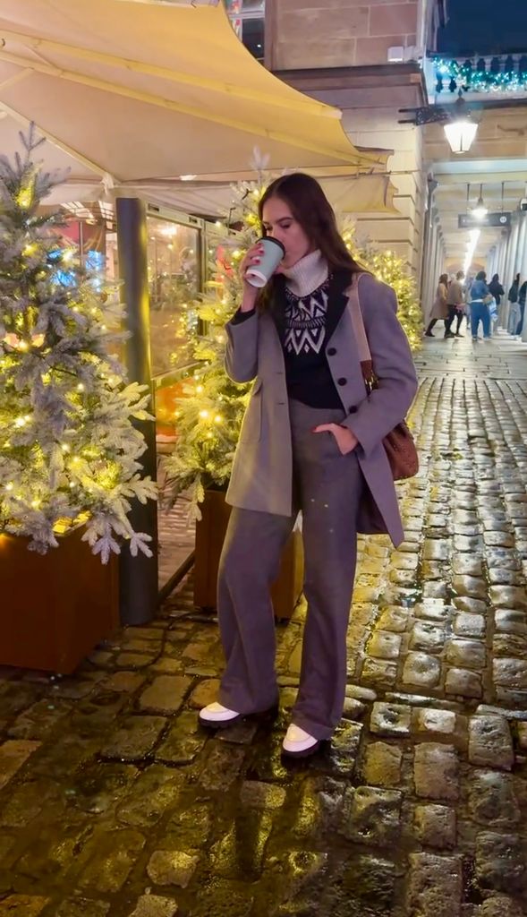 Natalie Salmon enjoys a hot drink outdoors, styled in Sezane trousers, a festive Fair Isle sweater, a tailored grey coat, and white sneakers. The backdrop features sparkling Christmas trees and cobbled streets, exuding cozy holiday vibes.