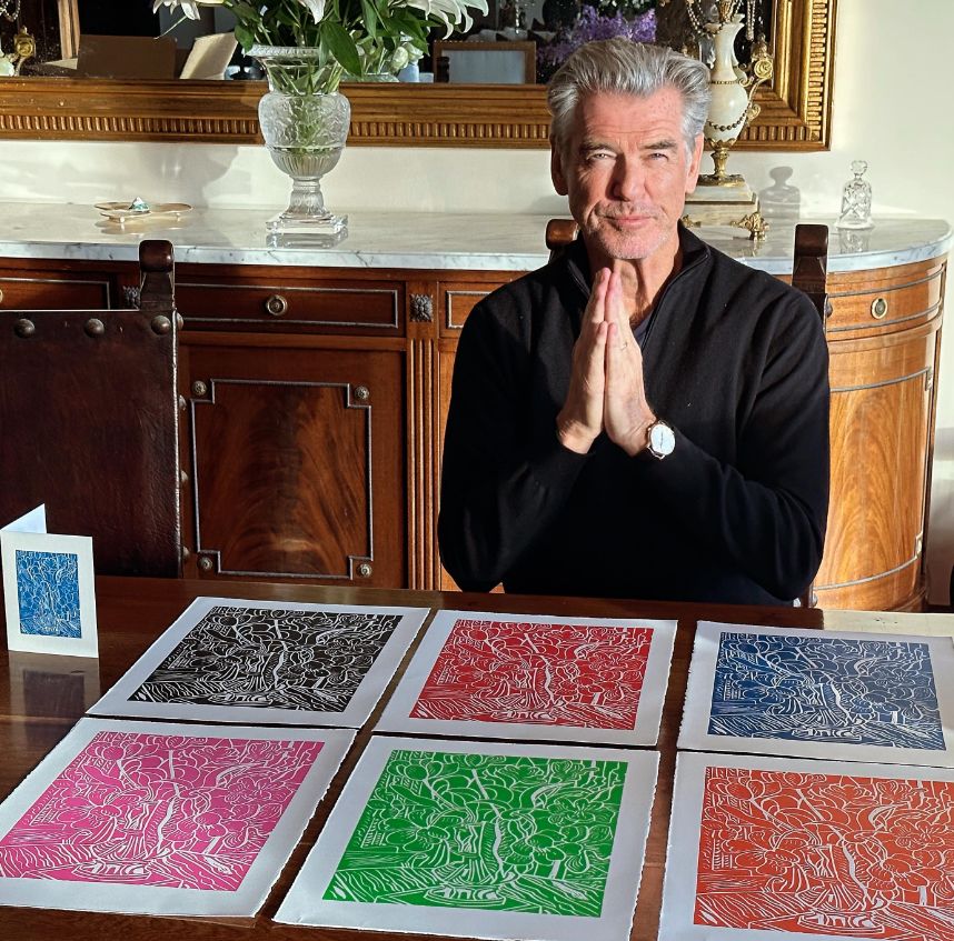 Pierce Brosnan sat at a table with six coloured prints in front of him