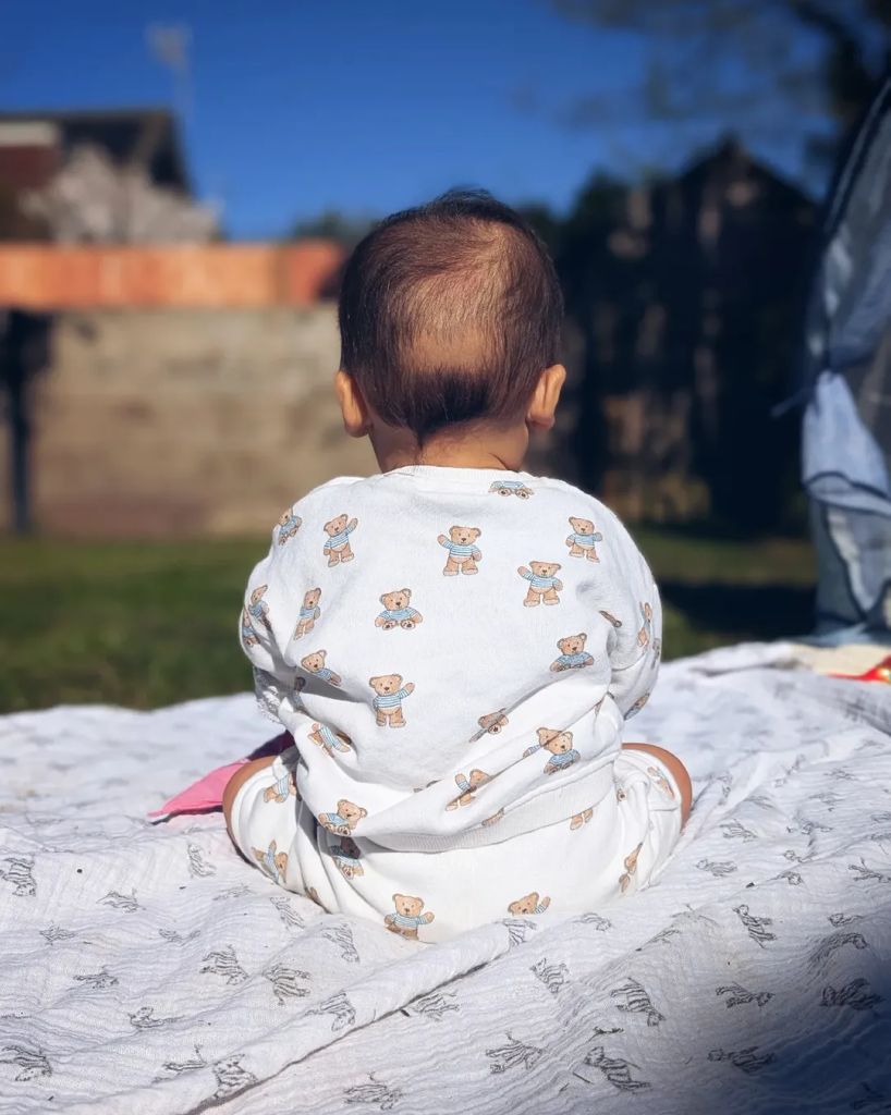 Will Kirk's son in a teddy-print outfit sitting in the garden with his back to the camera