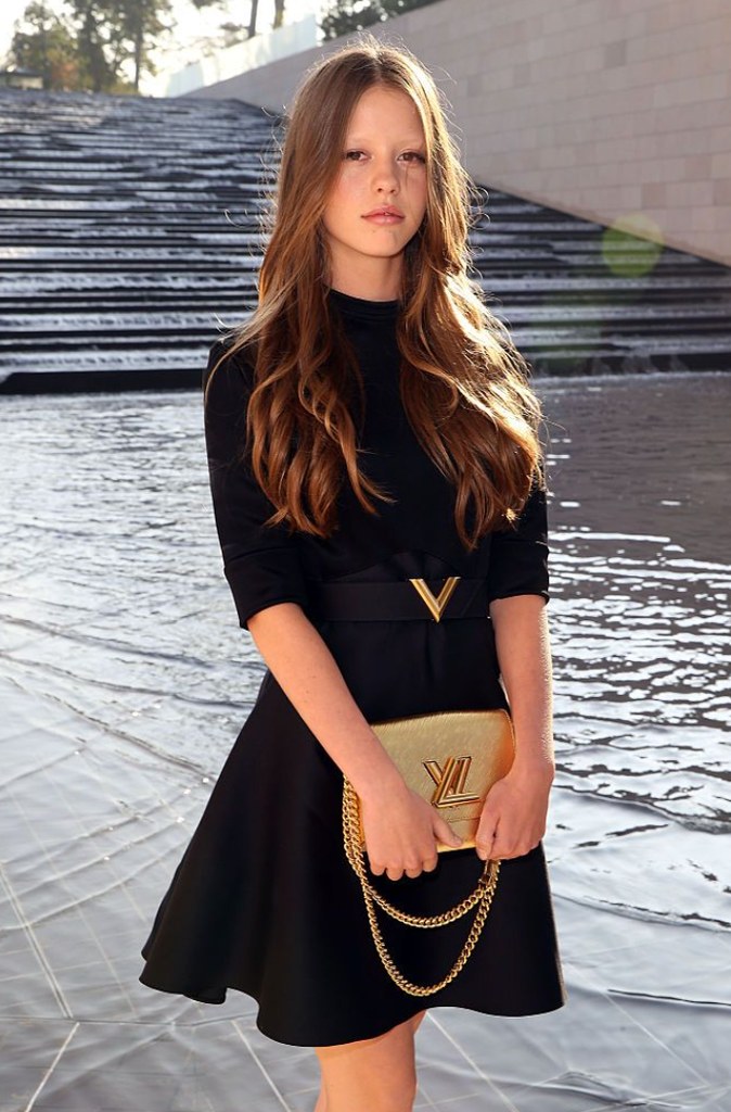 Mia Goth in 2015 wears a black mini dres with elbow length sleeves and gold belt, and holds a Louis Vuitton gold bag in front of her