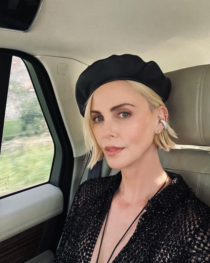 Charlize Theron in a black beret and dress sat in a car, smiling