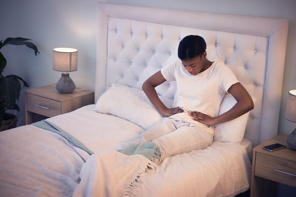 Pain, bed and woman with tummy cramps in her room holding her stomach for comfort. Tired, sick and African female person taking a nap with period ache for constipation or virus at her apartment.