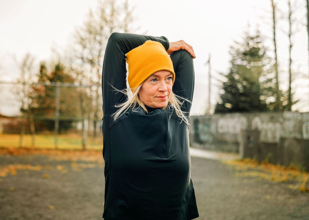 Mature woman stretching arms in the city park. Female jogger wearing knitted hat doing warm up workout outdoor on a winter morning.