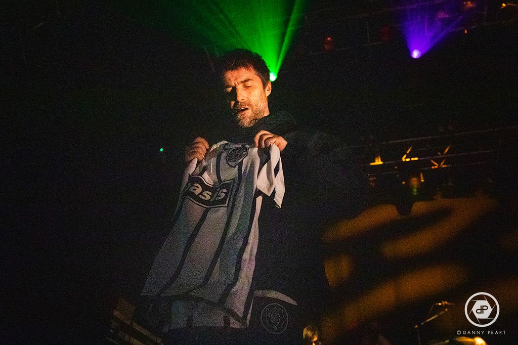 Liam Gallagher holding up football shirt on stage at gig
