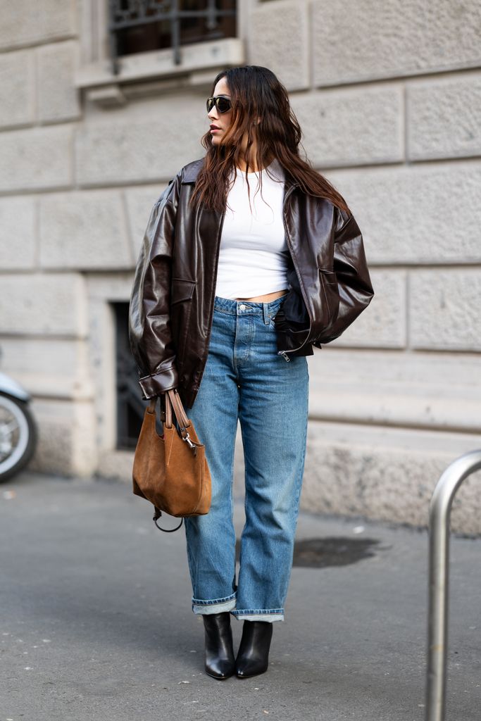 Teresanna Pugliese wears a dark brown leather bomber jacket over a white ribbed tank top and straight blue jeans, paired with black pointed boots, a brown suede handbag, and dark sunglasses, with loose wavy hair outside Pilot Room showroom on November 13, 2025 in Milan, Italy. (Photo by Valentina Frugiuele/Getty Images)