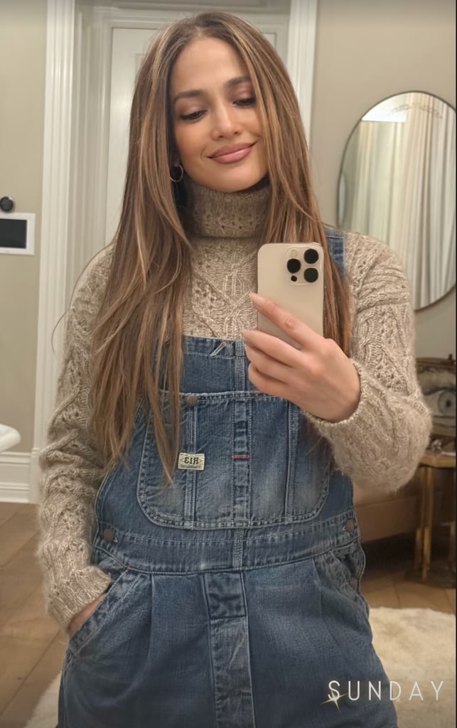 Jennifer Lopez standing in a warmly lit, neutral-toned room, taking a mirror selfie. She wears blue denim dungarees over a cozy beige knit sweater, her long highlighted hair framing her serene face. A round mirror and a traditional wooden console table are faintly visible in the background, enhancing the cozy, sophisticated atmosphere. The word "SUNDAY" appears discreetly in the corner of the photo.