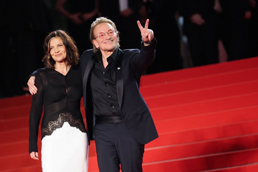Ali Hewson et Bono se tiennent ensemble sur le tapis rouge, Bono brandit un signe de paix et a un bras autour des épaules de sa femme.