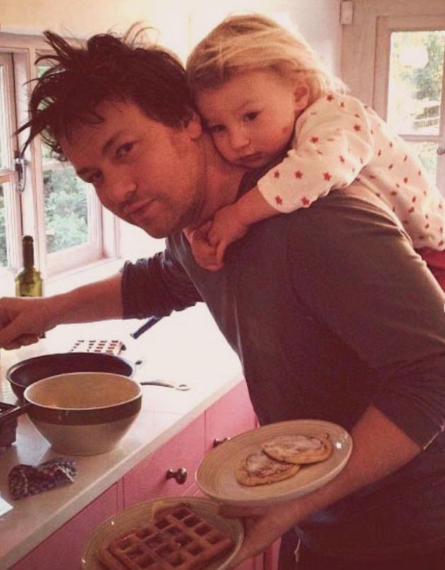 Jamie Oliver with a baby on his back balancing plates on his arm