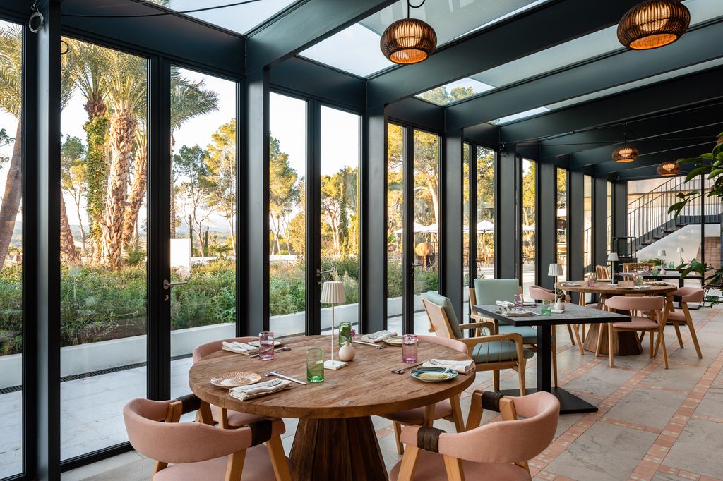 The Saba restaurant at Kimpton Aysla Mallorca, featuring stylish dining with panoramic glass walls that offer views of lush palm trees and scenic outdoor surroundings.