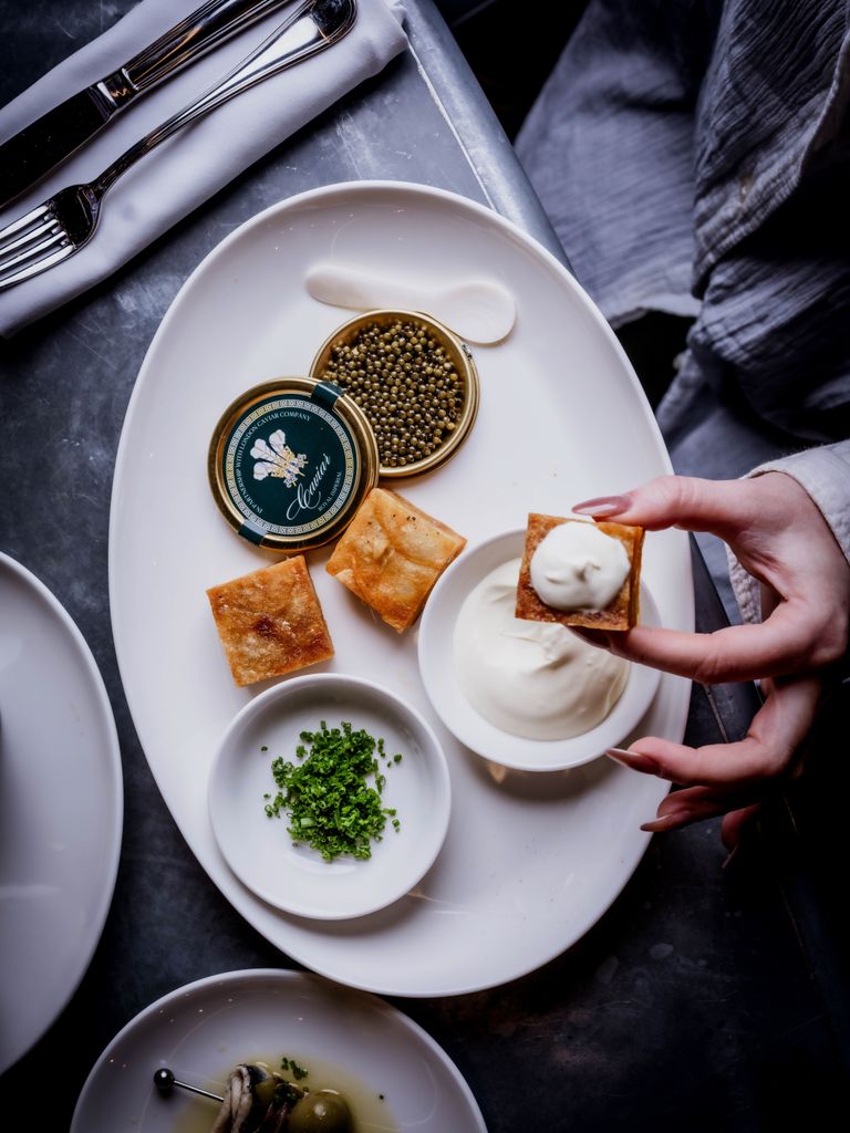 Caviar service at Prince Arthur pub, featuring a tin of sturgeon roe, blinis, crème fraîche, and chives, with a hand holding a blini.