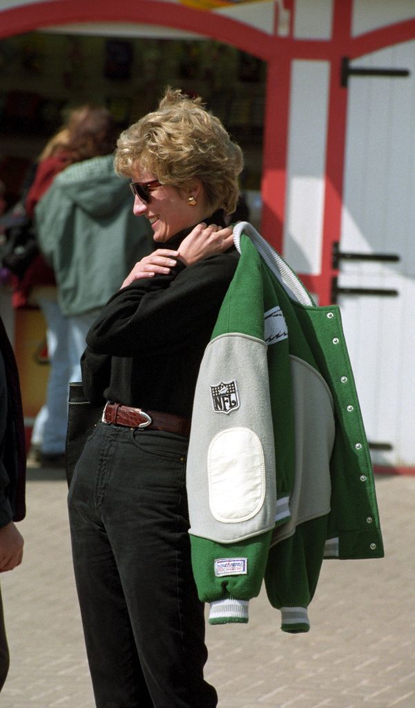 Princess Diana, glowing in the sun, slings her Philadelphia Eagles varsity jacket over her shoulder. Wearing a chic black top and sunglasses, she embodies ‘90s off-duty style at its finest.