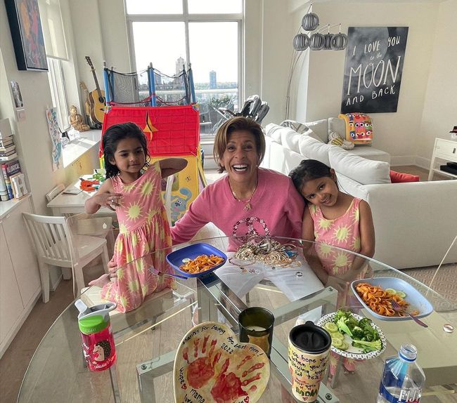 hoda sits between two little girls wearing matching pink dresses as they smile for the camera while seated at a round glass table filled with snacks and a handmade pot in a room filled with toys and a stunning view of new york city is visible from the window behind them