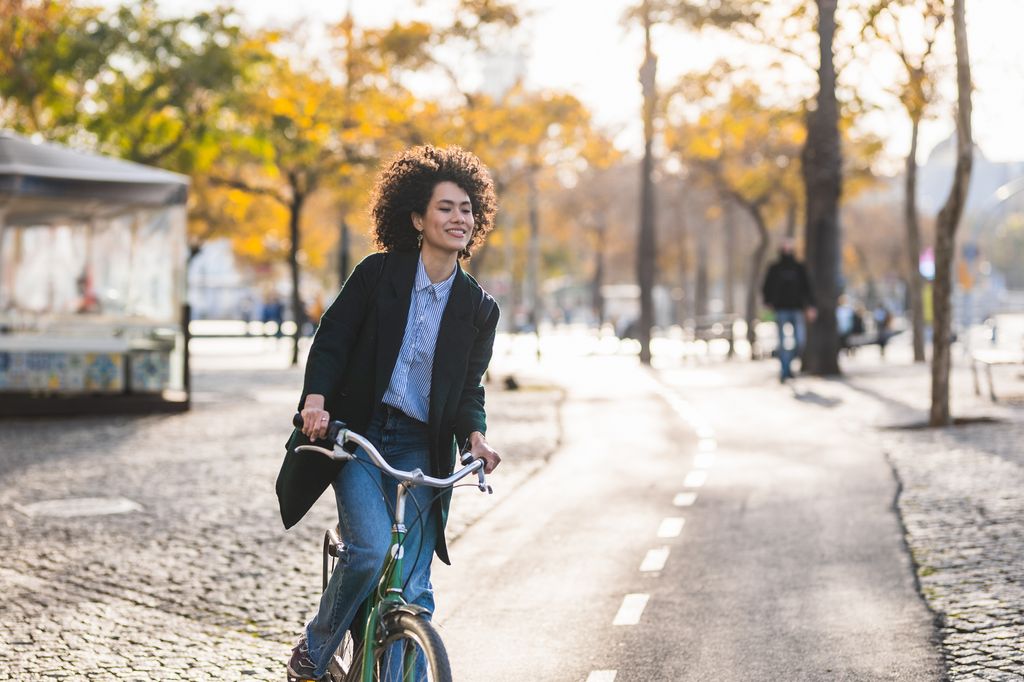 Beautiful woman with modern and relaxed look riding bike through park in autumn. Smiling happily while feeling the wind on her face, trees with orange leaves in the background.