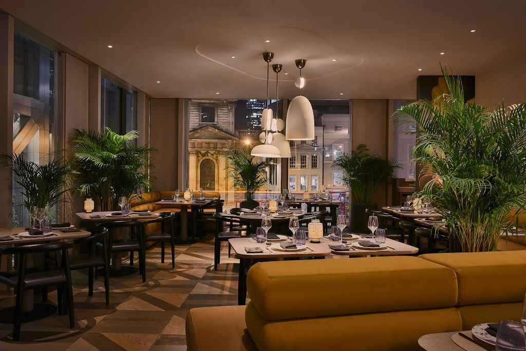 A warm and inviting image of the Straits Kitchen restaurant at The Pan Pacific London hotel, featuring set tables, lush potted plants, and a view of the city at night.