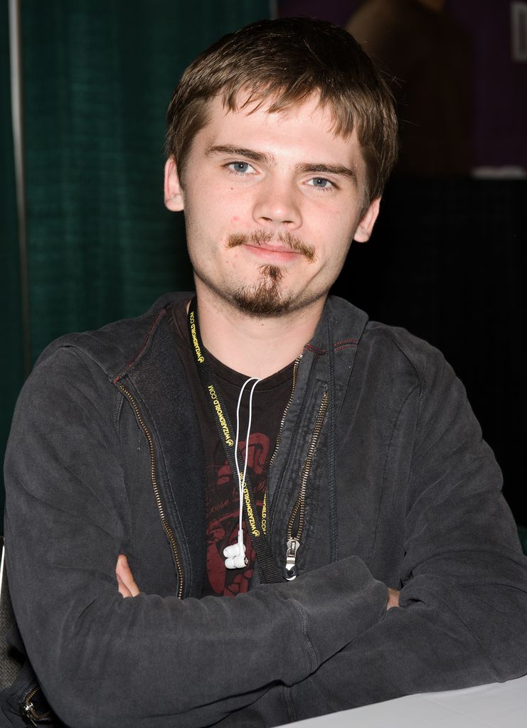 Jake Lloyd as a teeenager with a goatee and short brown hair, wearing a grey sweater