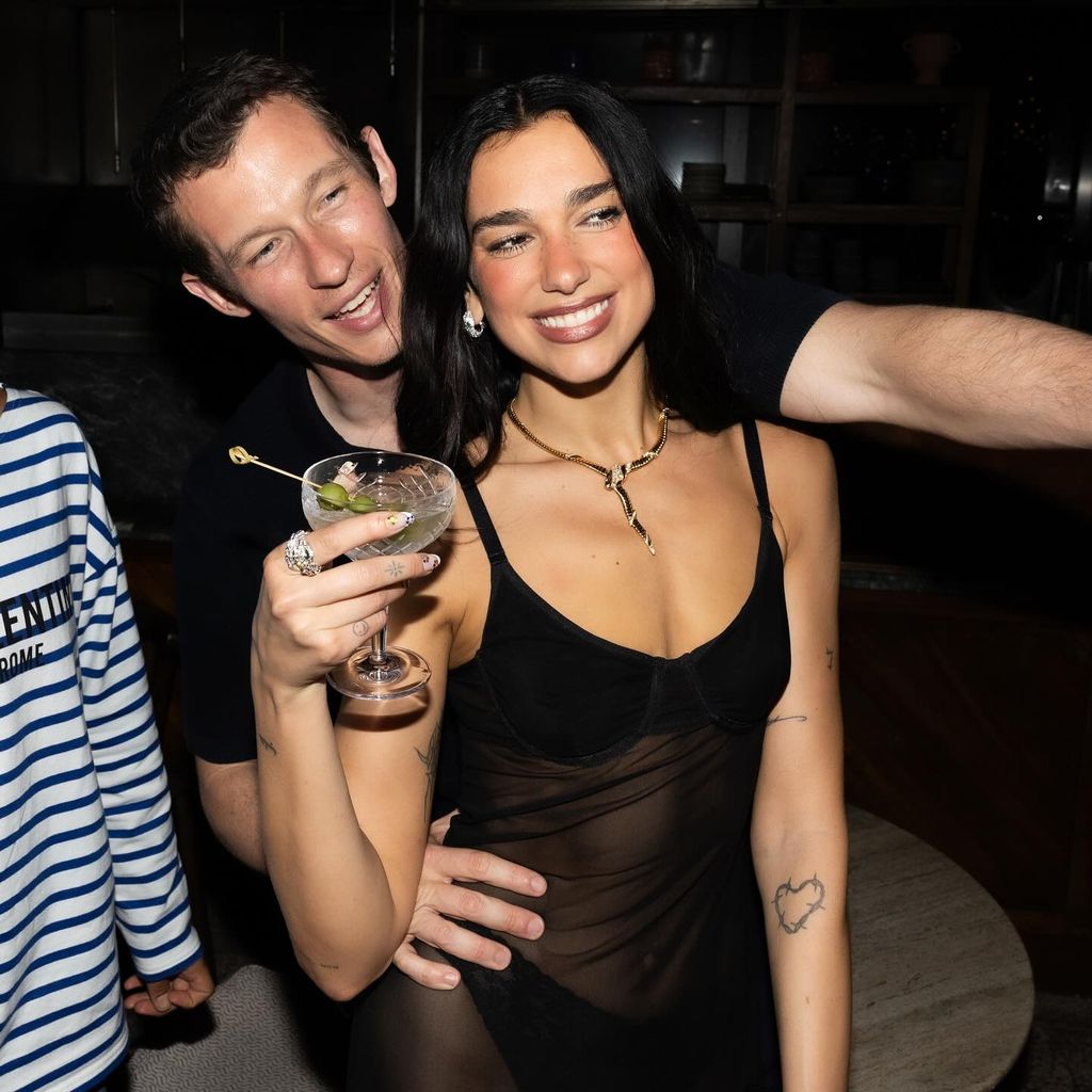 Dua Lipa and fiancé Callum Turner, Dua wears a sheer black dress and holds a martini glass, Callum has one hand on her hip and is wearing a black tshirt