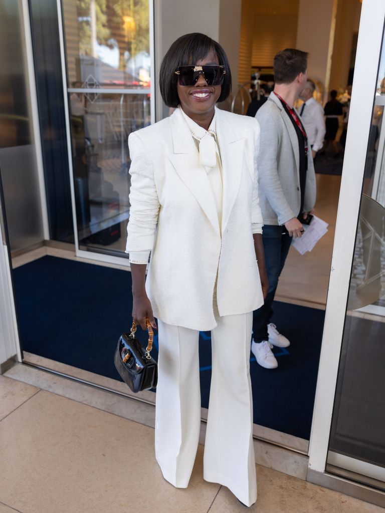 Viola Davis wears a white suit jacket, wide leg trousers and matching shirt, with black accessories - wide sunglasses and a top handle bag