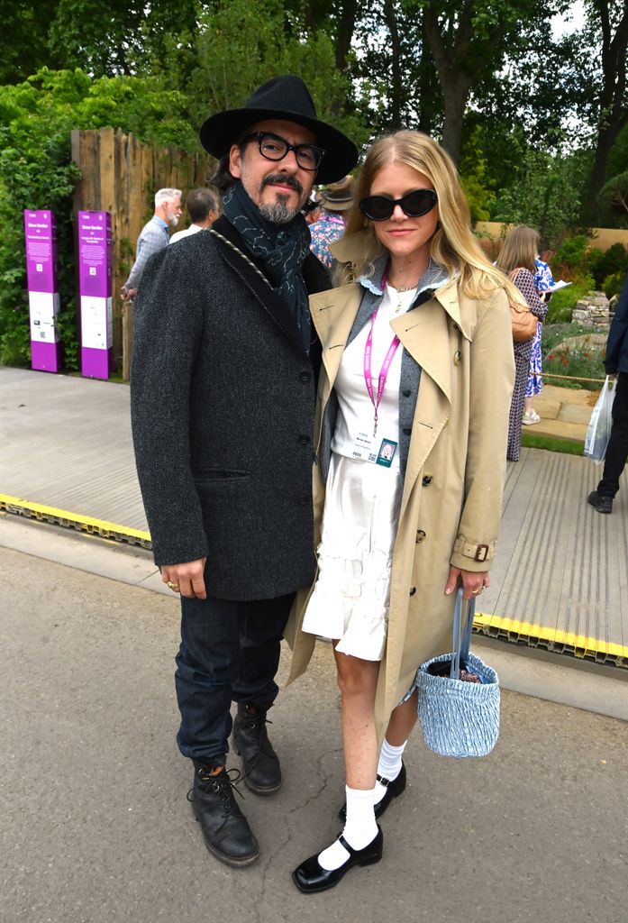 Dhani Harrison in a black coat and hat stands beside Mereki Beach in a trench coat and white dress with Mary Janes at Chelsea.