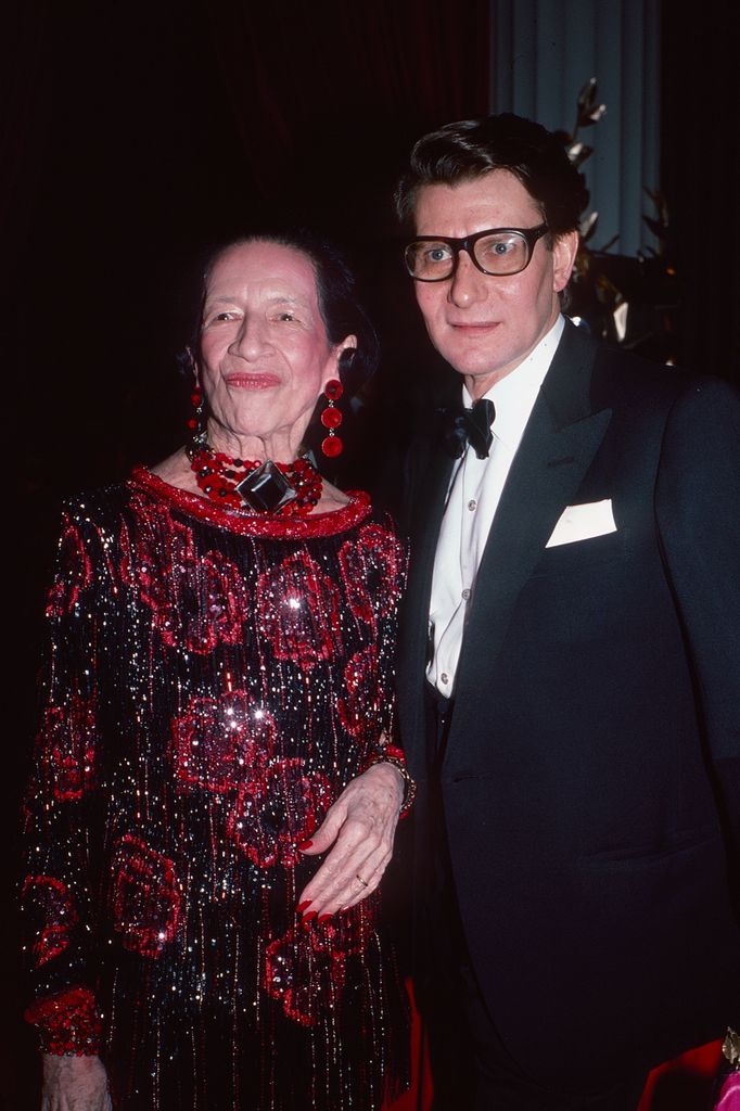 Diana Vreeland poses in a glittering red dress with bold red statement jewelry alongside Yves Saint Laurent at a Met Gala, channeling her signature theatrical elegance and editorial intensity.
