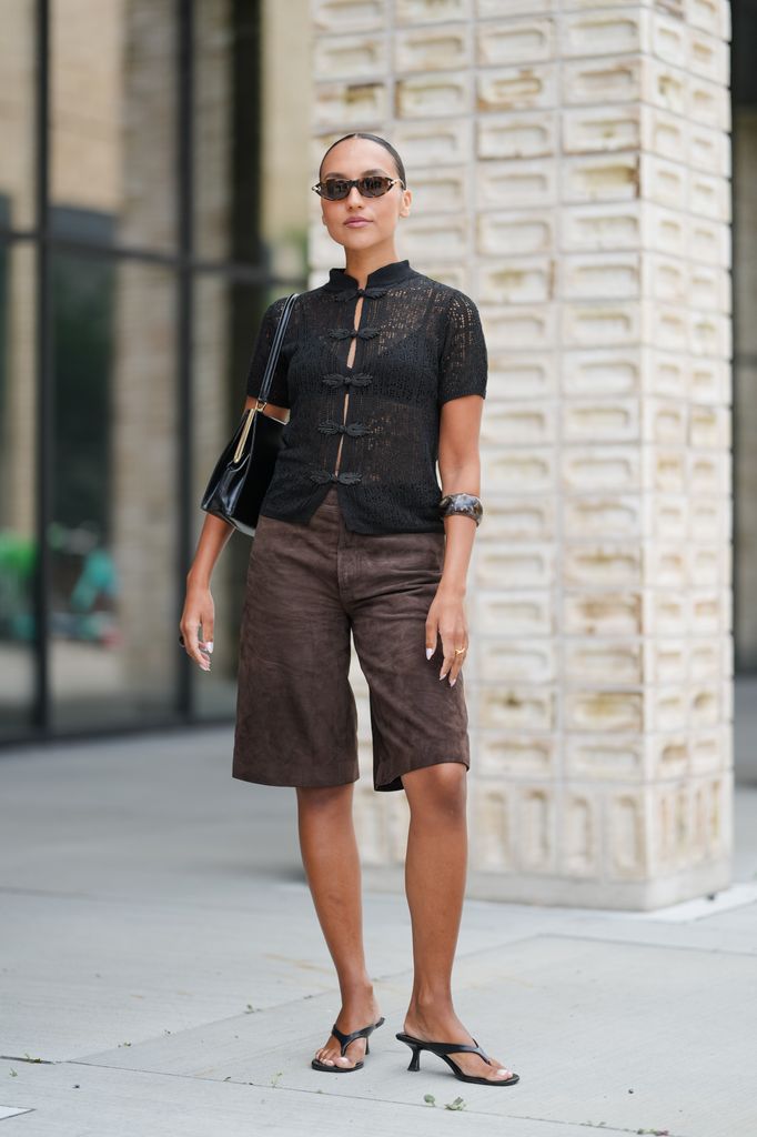 A guest wears a black, short-sleeved, sheer knit top with a high collar and decorative frog closures down the front. The top is paired with knee-length, brown suede shorts. Accessories include narrow, rectangular sunglasses with dark lenses, a black varnished leather shoulder bag with gold hardware, and a large, dark bangle on one wrist. 