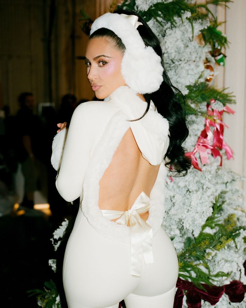 Kim Kardashian wearing a backless white catsuit and big fluffy ear muffs poses in front of a large green Christmas tree decorated with fake snow and pink bows