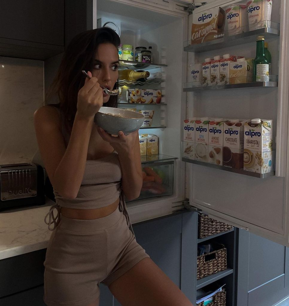 Lucy Watson comendo uma tigela de comida em frente à geladeira