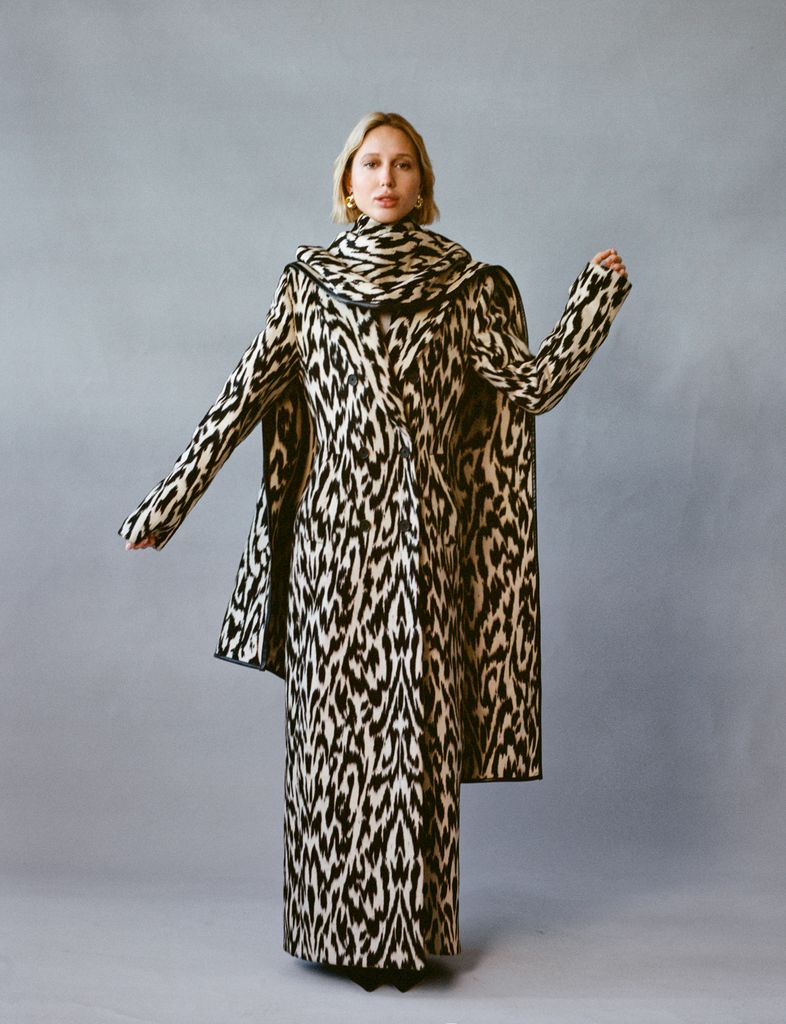 Princess Olympia of Greece showcases a patterned coat, extending one arm slightly to highlight its dramatic silhouette. The coordinating scarf frames her shoulders, adding to the bold, statement-making effect of the ensemble.
