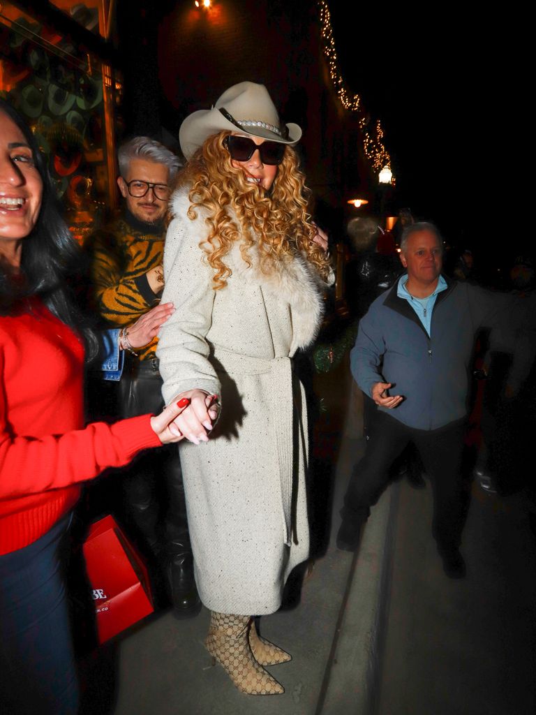 Mariah Carey in Colorado wearing a cowboy hat, long coat and boots. She holds the hand of a woman in a red jumper and smiles at the camera wearing sunglasses