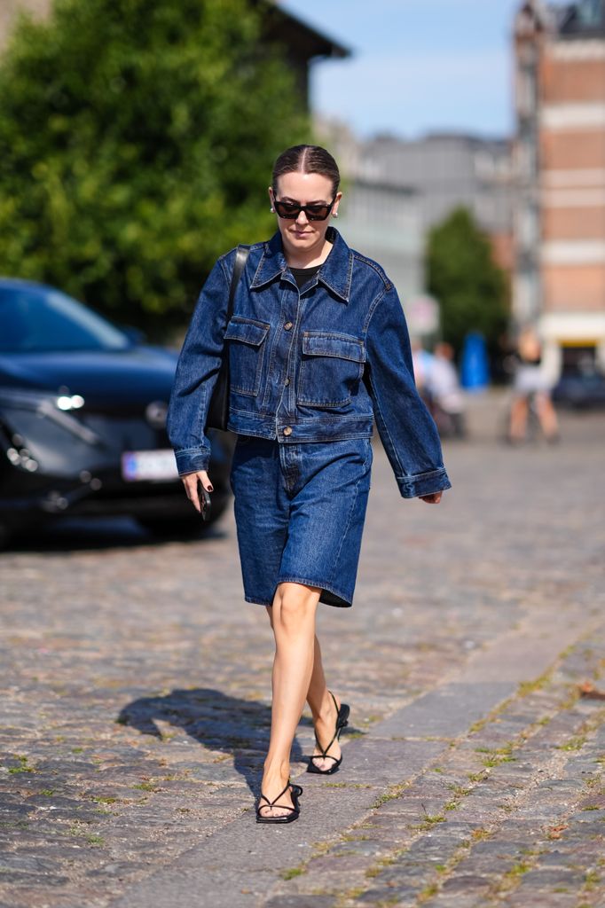 : A guest wears black sunglasses, silver earring, black t-shirt, dark navy blue denim jean jacket, dark navy blue denim jean shorts, black bag, strappy open toed leather kitten heels, outside Herskind, during the Copenhagen Fashion Week Spring/Summer 2024-2025 