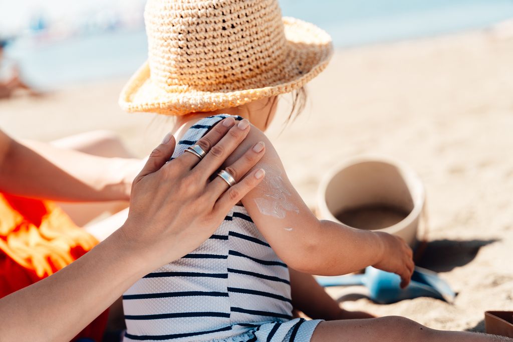 Asian mother putting sunscreen lotion on her daughterâs shoulders while sunbathing on the beach. Summer vacation.